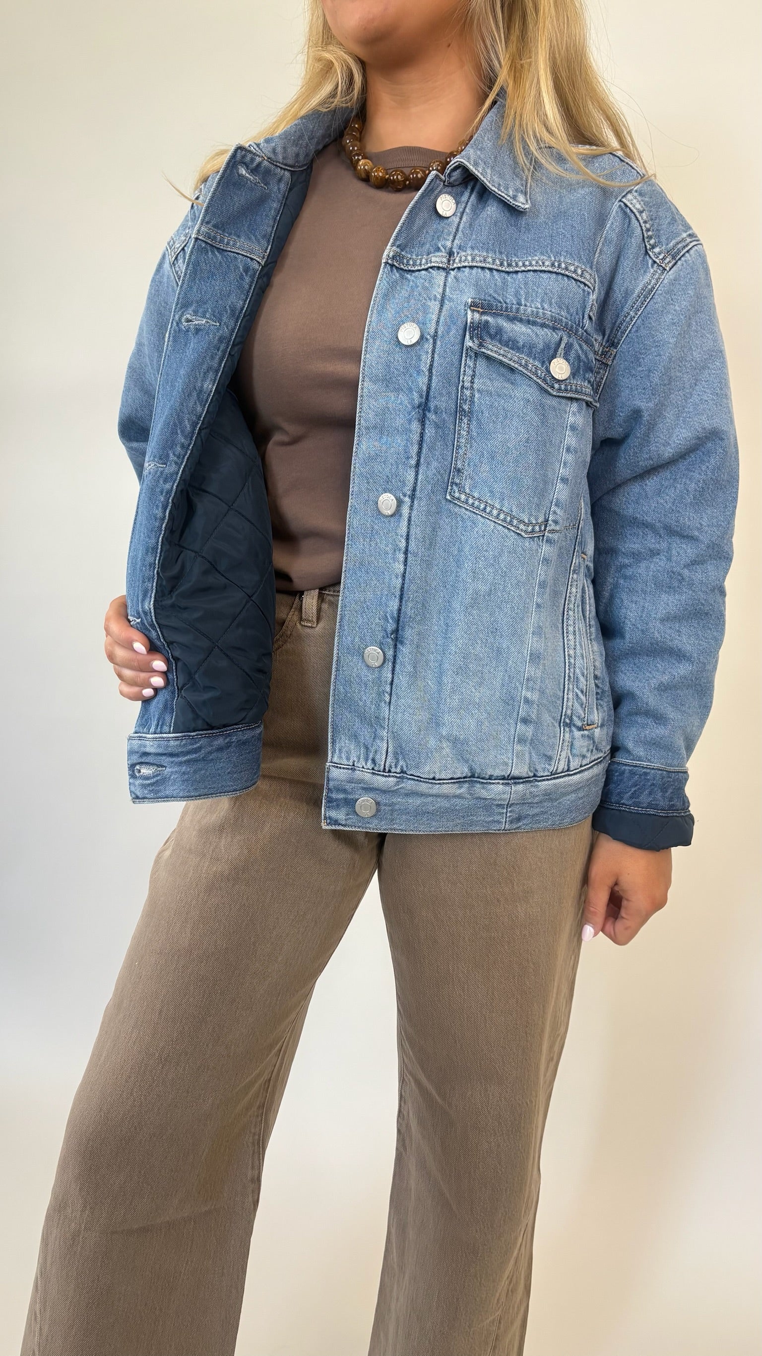 Person wearing a blue denim jacket over a brown shirt and pants on a plain background