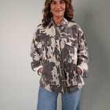 A woman wearing a gray and white abstract patterned zip-up jacket and blue jeans, photographed against a plain light gray studio background.