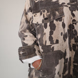 A close-up of a person wearing a distressed acid-wash denim jacket with a black and beige cow-print pattern, paired with blue jeans and a white undershirt.