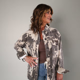 A woman modeling a brown and beige camouflage patterned jacket over a brown top and blue jeans, standing against a solid gray studio background.