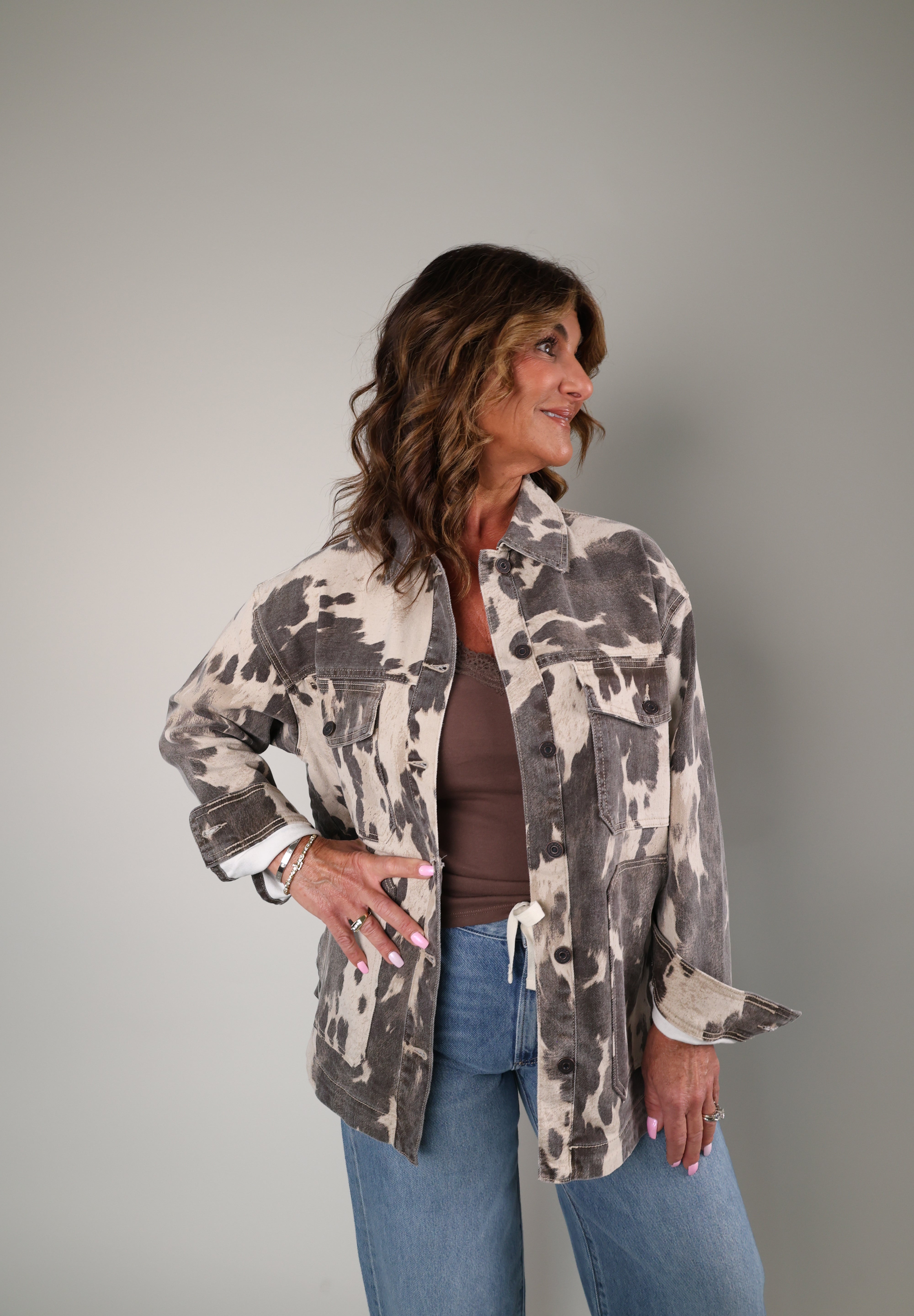 A woman modeling a brown and beige camouflage patterned jacket over a brown top and blue jeans, standing against a solid gray studio background.