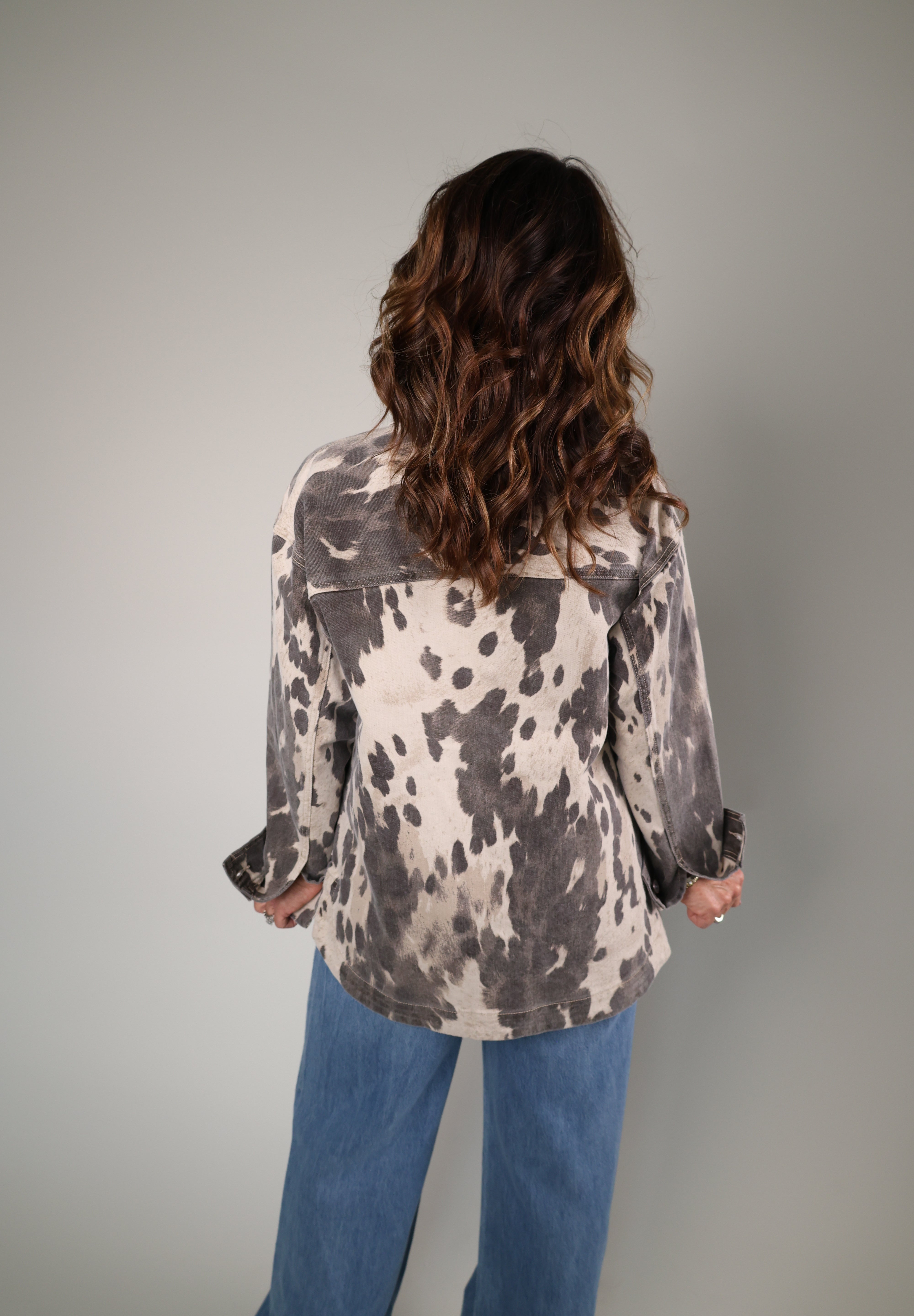 A woman viewed from behind wearing a long-sleeve button-up shirt with a black and white cow-print pattern, paired with blue jeans, against a plain light gray studio background.