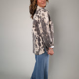 A woman modeling a long-sleeve button-up shirt with a gray and off-white tie-dye pattern, paired with wide-leg blue jeans, photographed against a neutral gray studio background.