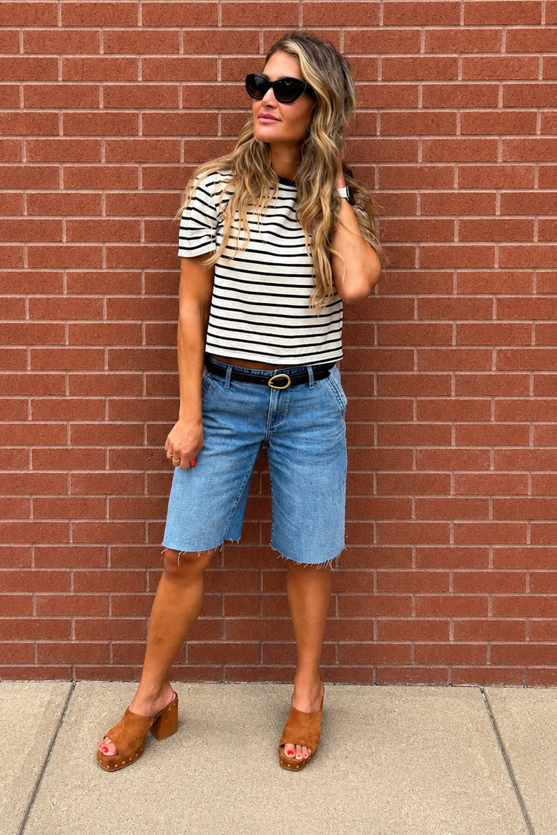 A woman stands against a red brick wall wearing a black and white striped short-sleeve t-shirt, light blue denim shorts, a black belt, and brown leather clogs.