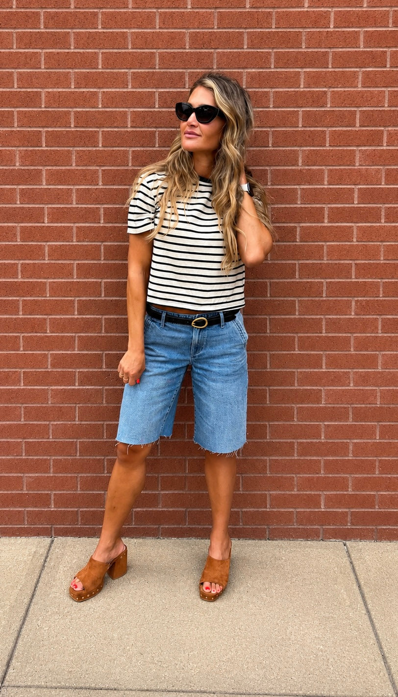 A woman stands against a red brick wall wearing a black and white striped short-sleeve t-shirt, light blue denim shorts, a black belt, and brown leather clogs.