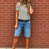 A woman stands against a red brick wall wearing a black and white striped short-sleeve t-shirt, light blue denim shorts, a black belt, and brown leather clogs.