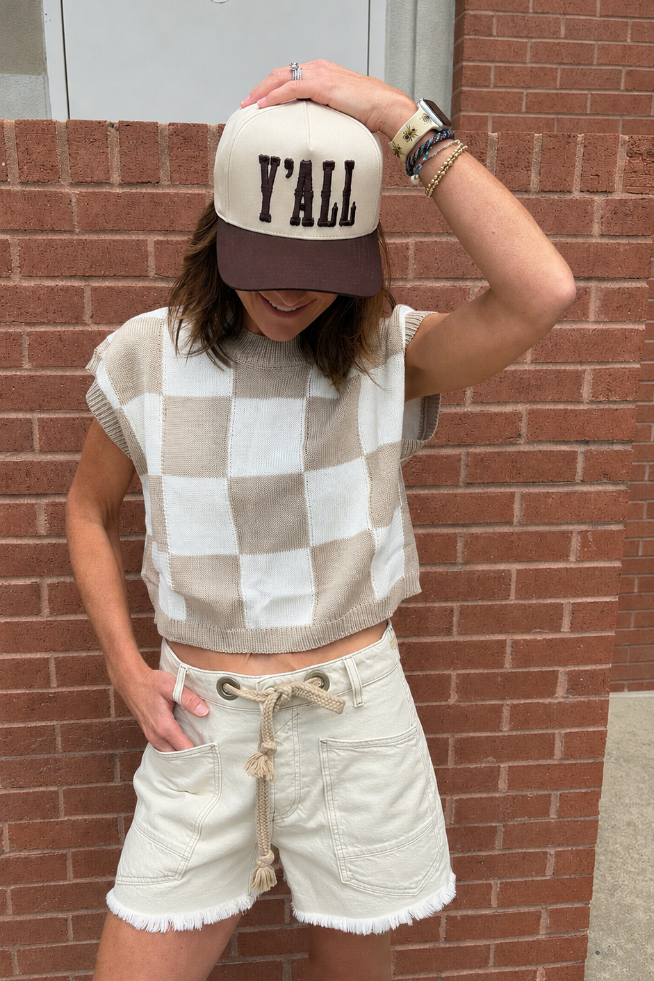 A woman wearing a beige and white checkered sleeveless knit crop top, cream frayed denim shorts, and a cream and brown baseball cap with 'Y'ALL' printed on the front, posing against a red brick wall.