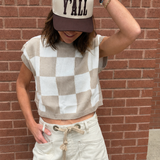 A woman wearing a beige and white checkered sleeveless knit crop top, cream frayed denim shorts, and a cream and brown baseball cap with 'Y'ALL' printed on the front, posing against a red brick wall.