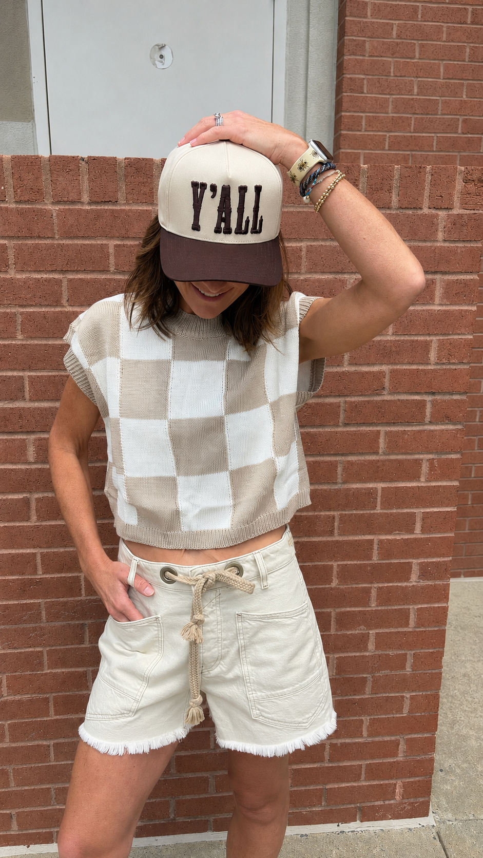 A woman wearing a beige and white checkered sleeveless knit crop top, cream frayed denim shorts, and a cream and brown baseball cap with 'Y'ALL' printed on the front, posing against a red brick wall.