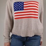 Woman wearing the Z Supply Sienna Patriot Sweater in a soft neutral knit with an American flag graphic on the front and denim shorts.