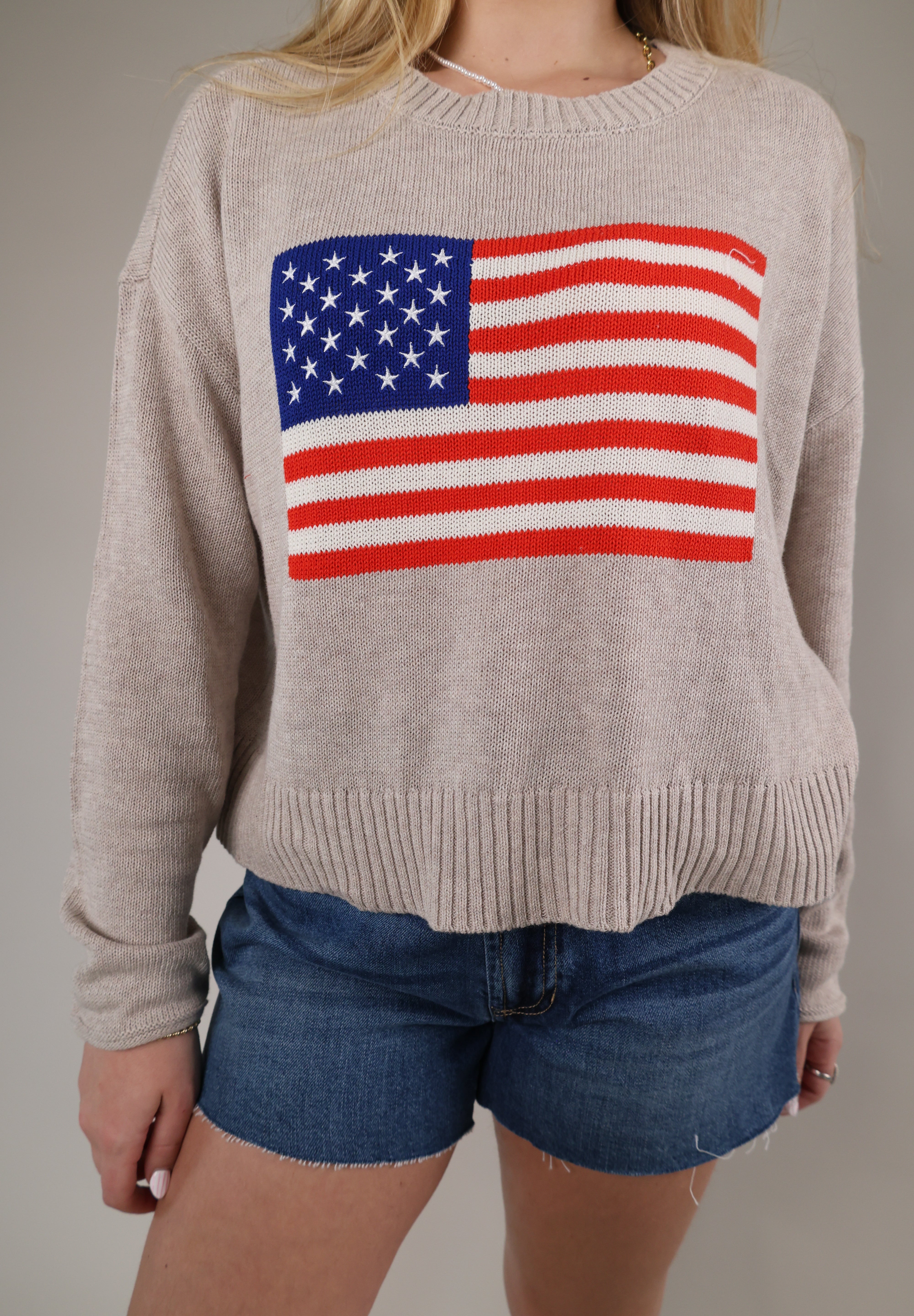 Woman wearing the Z Supply Sienna Patriot Sweater in a soft neutral knit with an American flag graphic on the front and denim shorts.